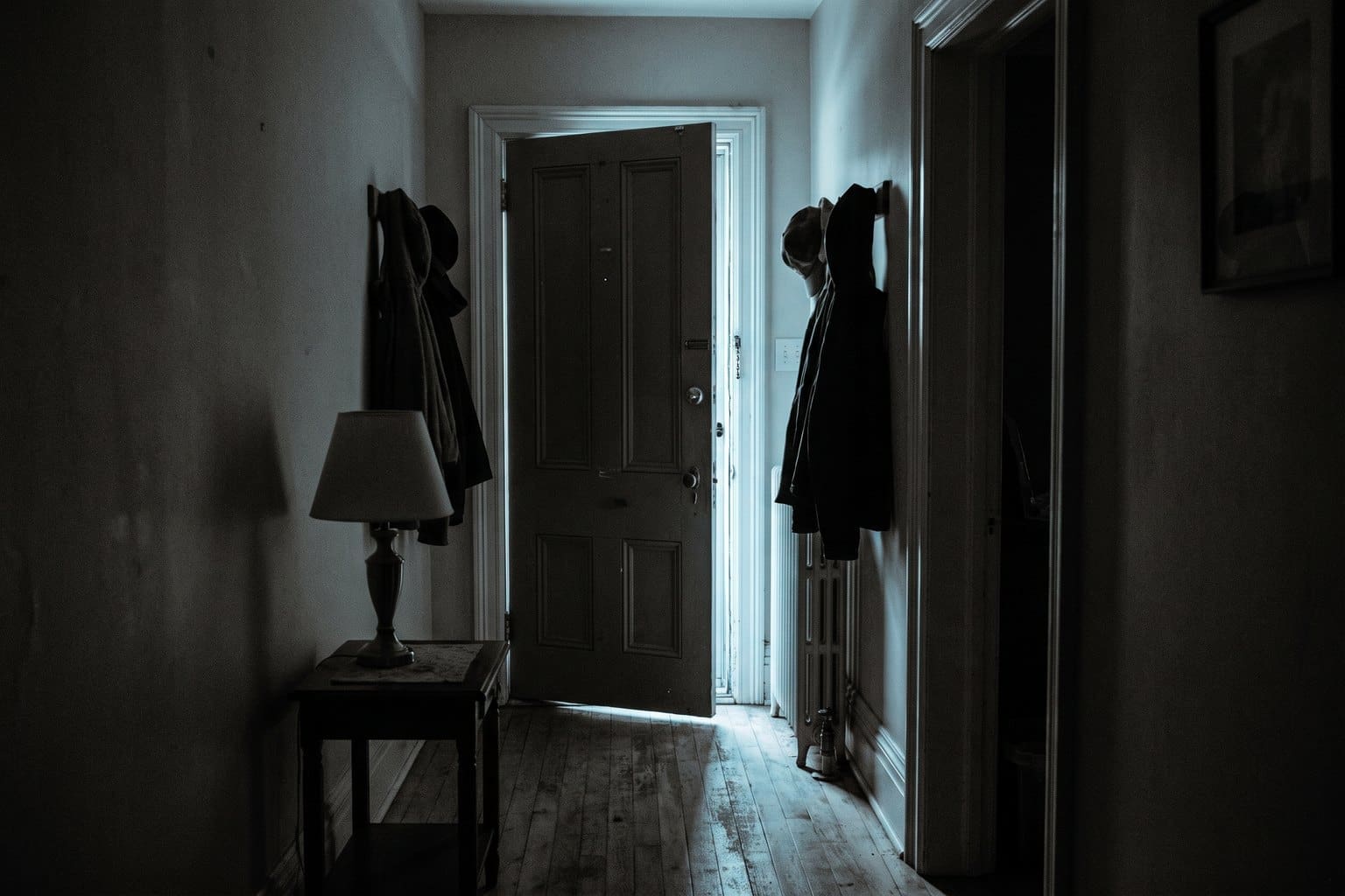 Domestic hallway at night seen from inside with the front door slightly ajar and cold light through the gap